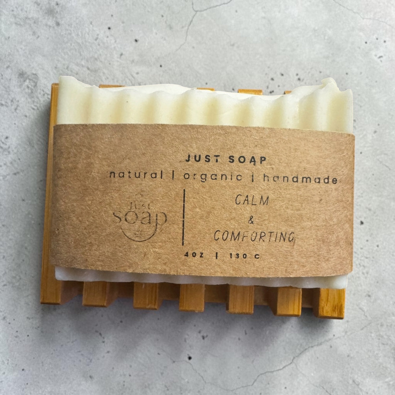 Just Soap Calm & Comforting handmade soap bar with kraft paper label on a bamboo soap dish, concrete background